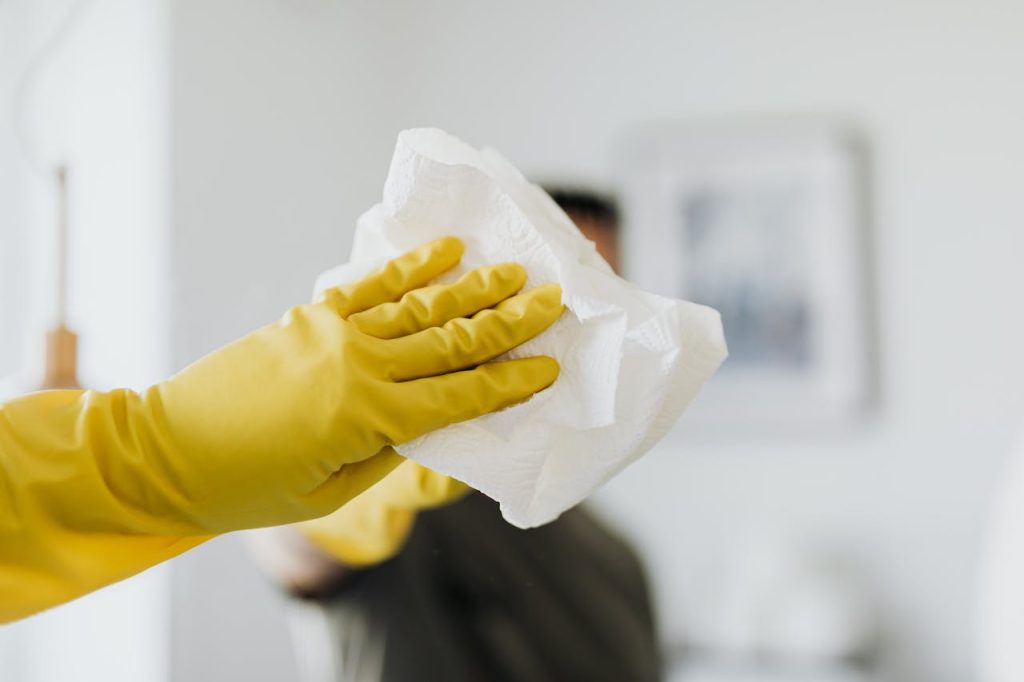 unrecognizable-man-cleaning-mirror-in-modern-bathroom-4239127 Close-up of a yellow-gloved hand cleaning a mirror with a cloth indoors.